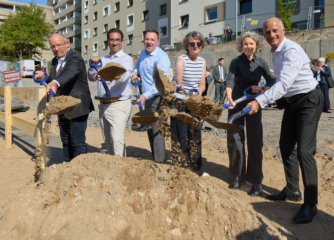Vier Männer und zwei Frauen, darunter die Ministerin Scharrenbach halten Schaufeln in der Hand und schippen Sand. Dabei lächeln sie in die Kamera.