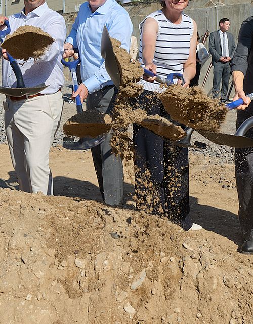 Vier Männer und zwei Frauen, darunter die Ministerin Scharrenbach halten Schaufeln in der Hand und schippen Sand. Dabei lächeln sie in die Kamera.