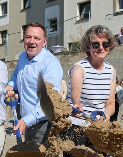 Vier Männer und zwei Frauen, darunter die Ministerin Scharrenbach halten Schaufeln in der Hand und schippen Sand. Dabei lächeln sie in die Kamera.