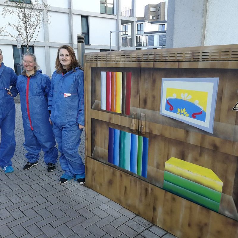 Studentinnen in Blaumännern vor dem besprühten Kasten