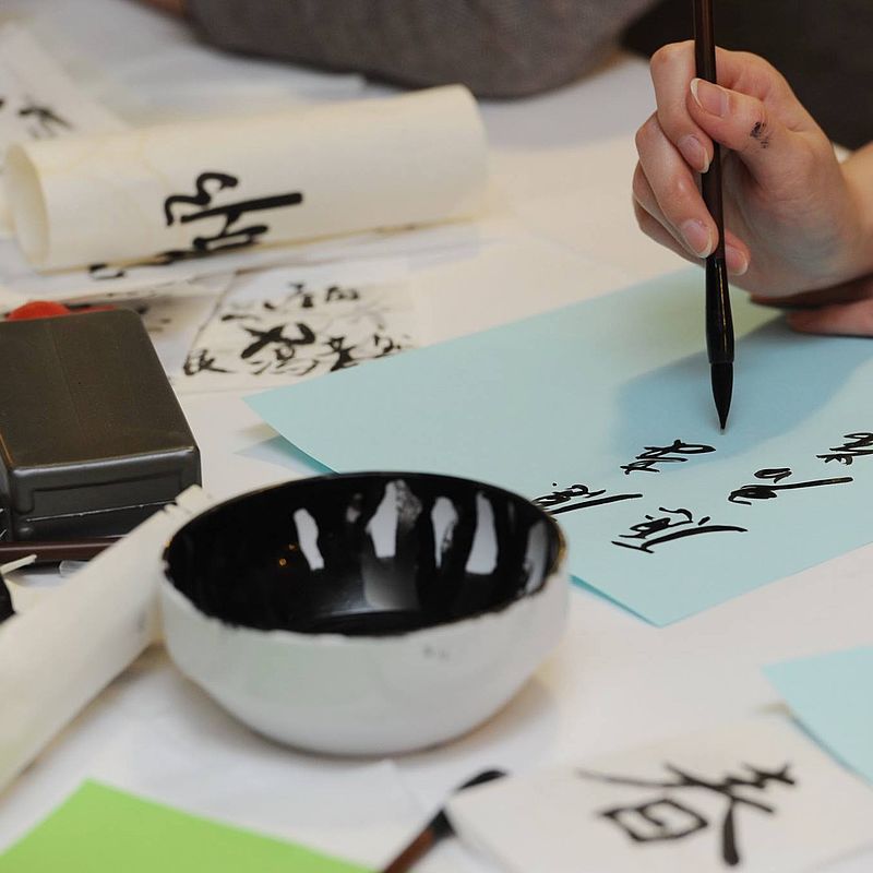 Schale mit schwarzer Tinte und eine Hand, die einen Pinsel in der Hand hält und chinesische Buchstaben aufs Papier bringt