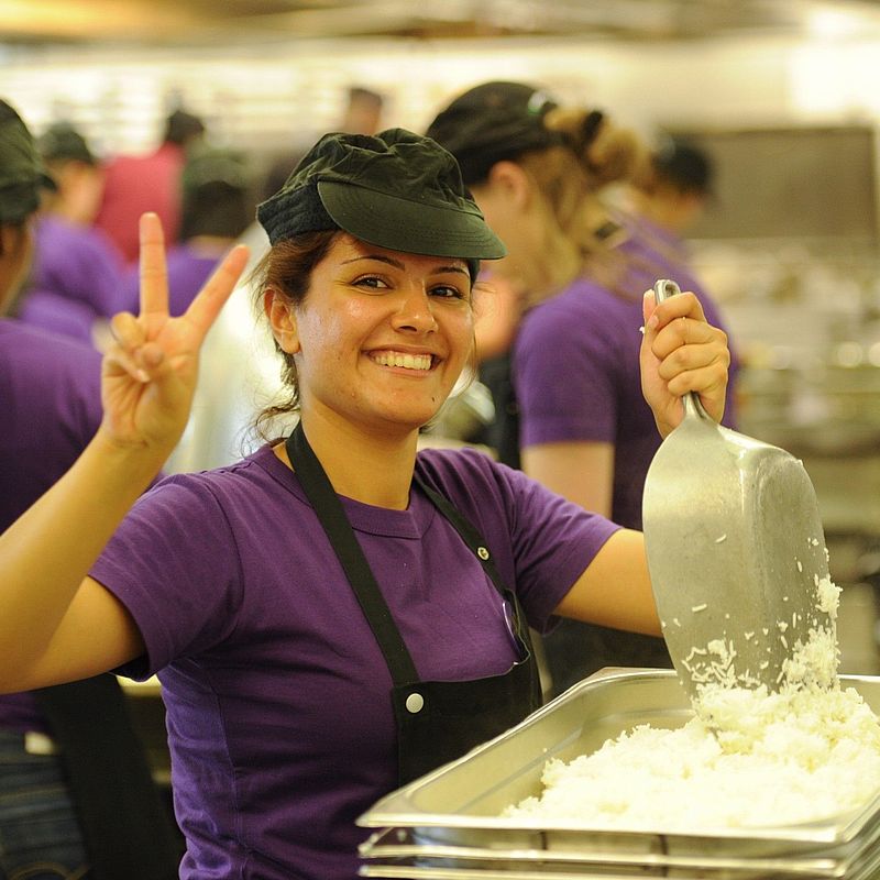 Studierende kochen beim Fest namens Culinara Festo in der Mensa-Küche, eine Studentin lächelt in die Kamera und zeigt das Peace-Zeichen