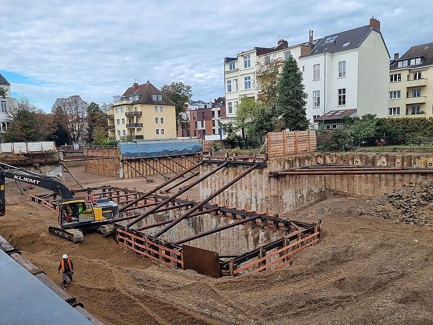 Städtische Baugrube mit Bagger und schrägen Stahlstreben, die Böschungen abstützen, Arbeiter und umliegende Wohnhäuser