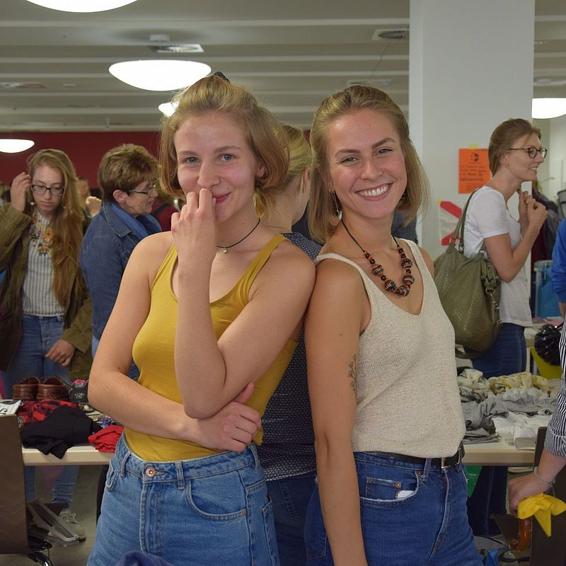 zwei Studentinnen posieren fürs Foto, im Hintergrund sieht man Kleiderständer und Markt-Gäste