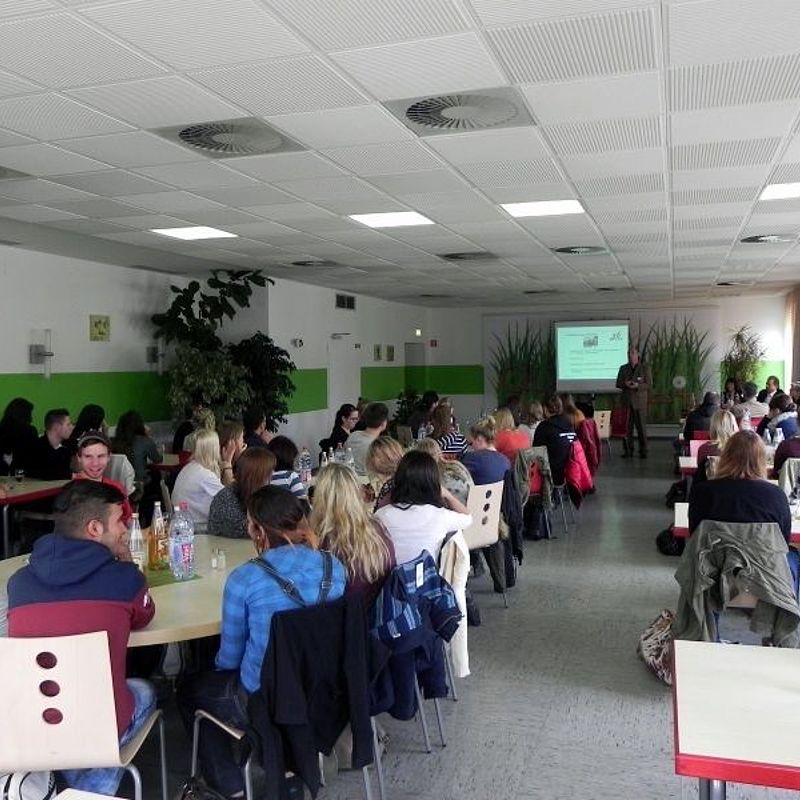 Mann steht vor einer Leinwand, auf die eine Präsentation projiziert wird und erklärt einem Saal voller Studierender etwas