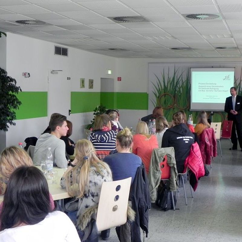 Mann steht vor einer Leinwand, auf die eine Präsentation projiziert wird und erklärt einem Saal voller Studierender etwas