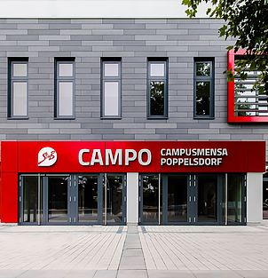 Zu sehen ist der Eingang der Mensa CAMPO. Das Gebäude ist dezent grau. Über der grauen Eingangstür prangert ein großes rotes Schild, das über die gesamte Türbreite reicht. Es ist mit "CAMPO Campusmensa Poppelsdorf" beschriftet.