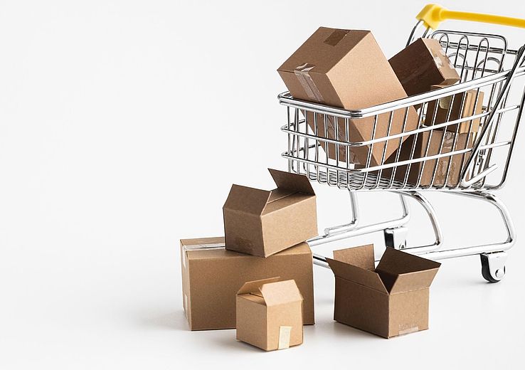 Shopping cart with a yellow handle filled with cardboard boxes and several boxes beside it on white background