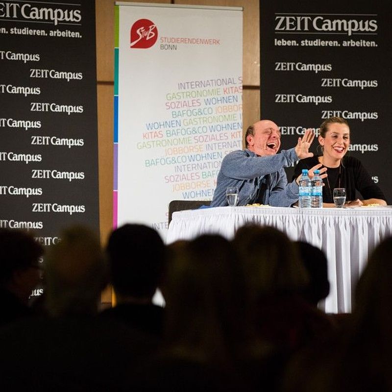 Bernhard Hoecker in der Mensa Nassestraße auf der Bühne an einem Tisch sitzend, zusammen mit der Moderatorin der ZEIT