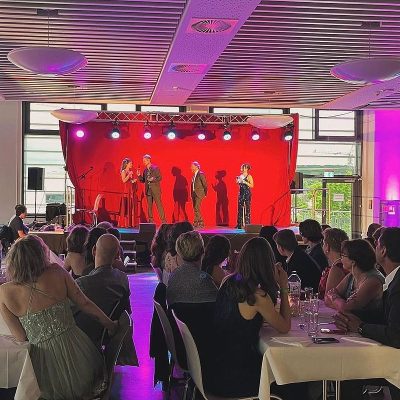 Perfekte Abi-Location in Bonn: Ein gefuellter feierlicher Saal mit Menschen, die an Tischen sitzen und nach vorne auf eine Buehne schauen, auf der eine Ehrung stattzufinden scheint. 