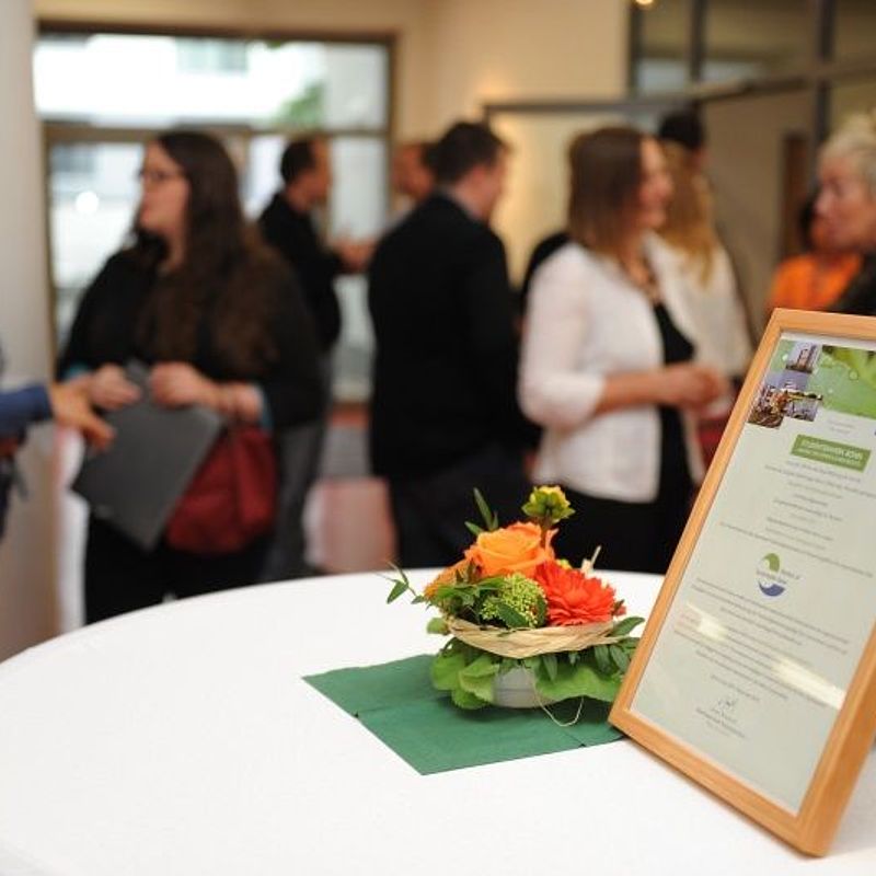 Urkunde Sustainable Bonn in einem Bilderrahmen auf einem Stehtisch mit Blumendeko