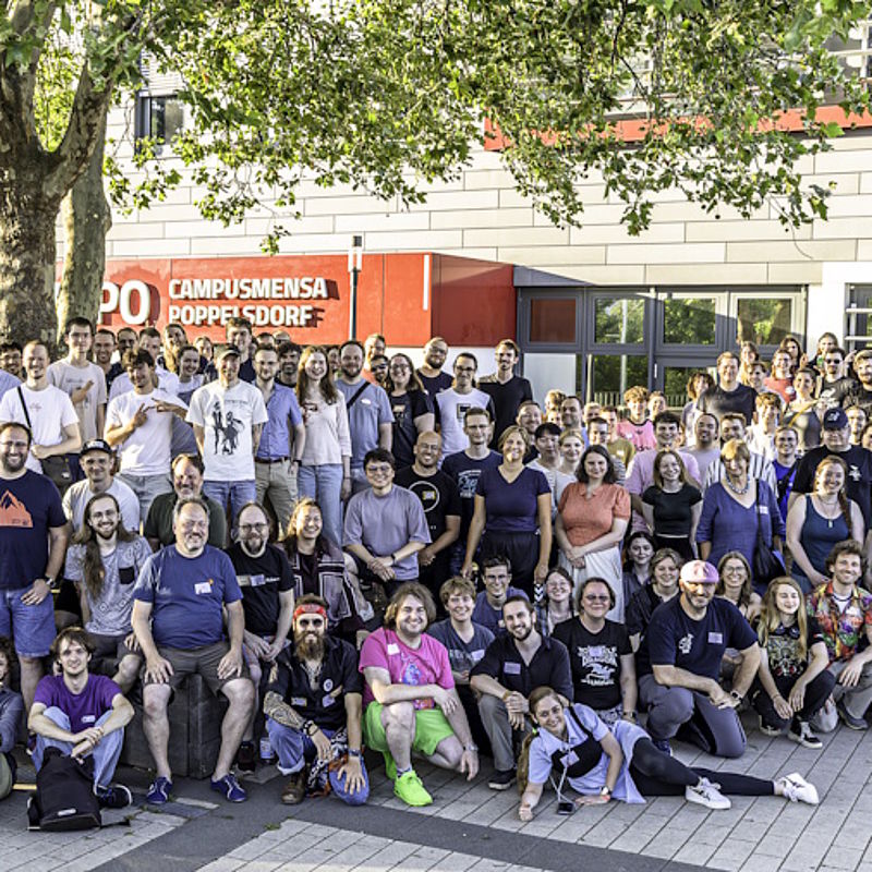 A group photo taken in front of the CAMPO entrance on a lovely summer’s day: all the guests and organisers are smiling as they gather around the tree, looking at the camera.