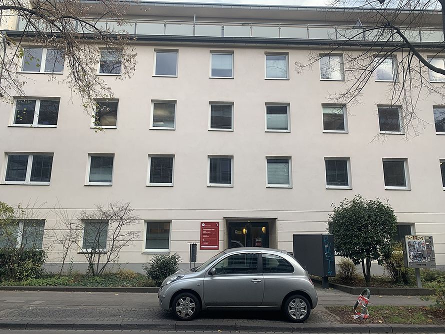 Fassadenfront eines Wohnhauses mit Reihen quadratischer Fenster, silbernes Auto parkend vor Eingang mit rotem Schild